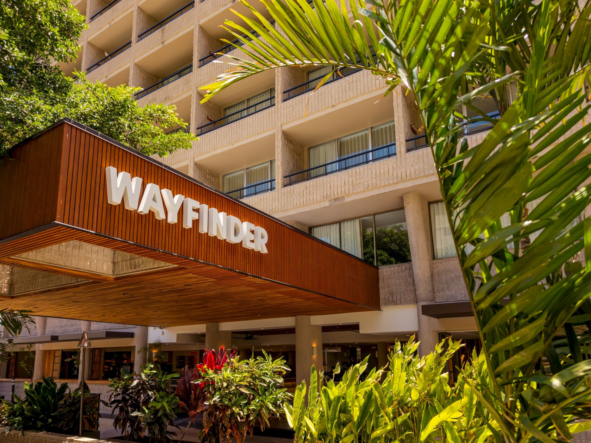 Wayfinder sign on a building entrance with tropical plants and a multi-story hotel or office behind it.