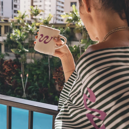 A person is standing on a balcony, holding a mug with the letter "w" on it, overlooking a pool and some apartment buildings.