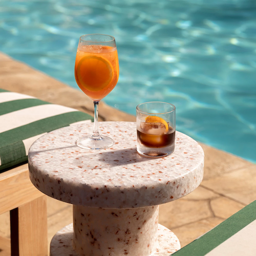 Two drinks on a small marble table next to a pool with striped cushions on the chairs.